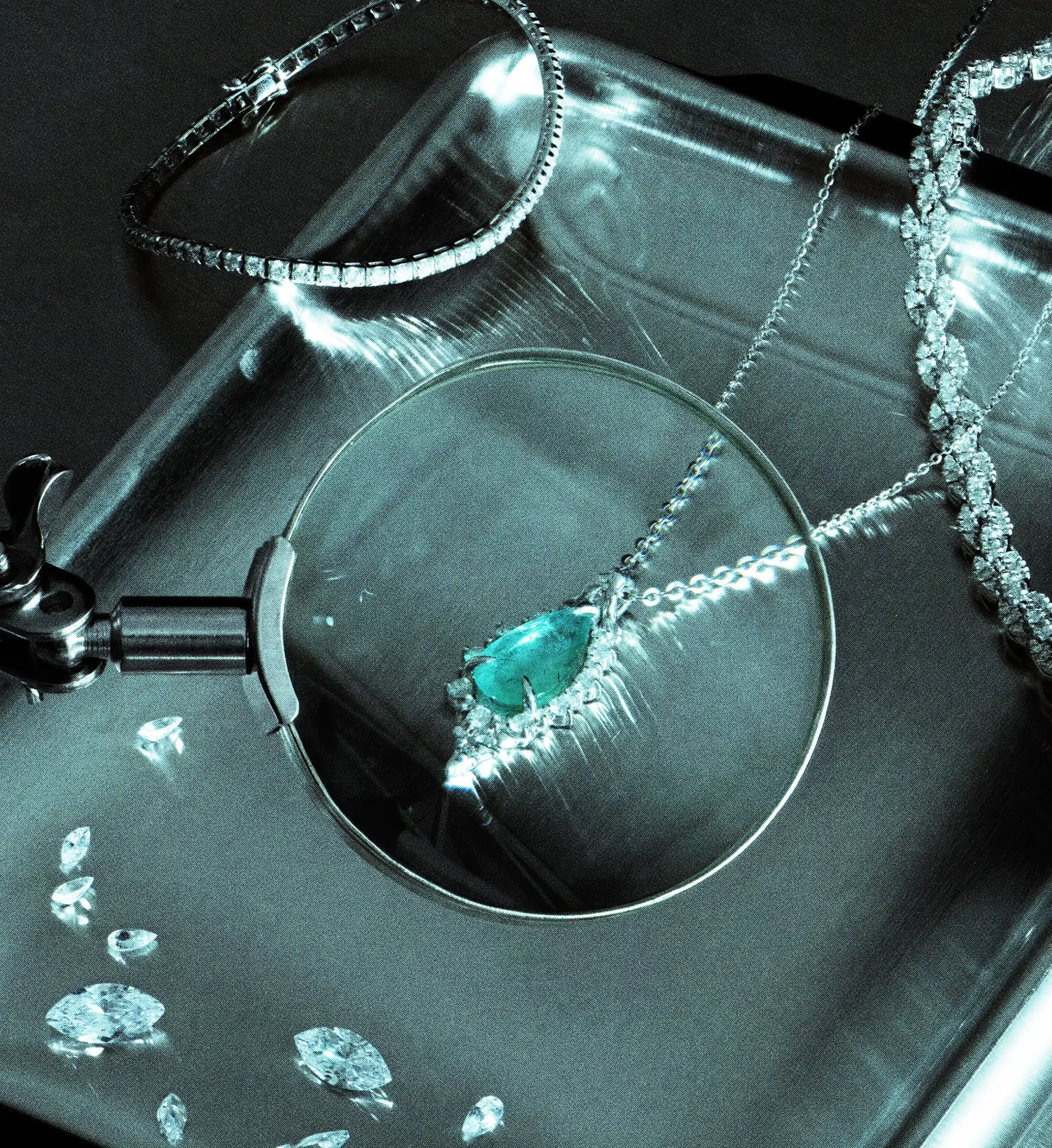 Pear-cut emerald pendant on a fine diamond chain, resting on a teal glass tray beside loose diamonds and a jeweller's loupe